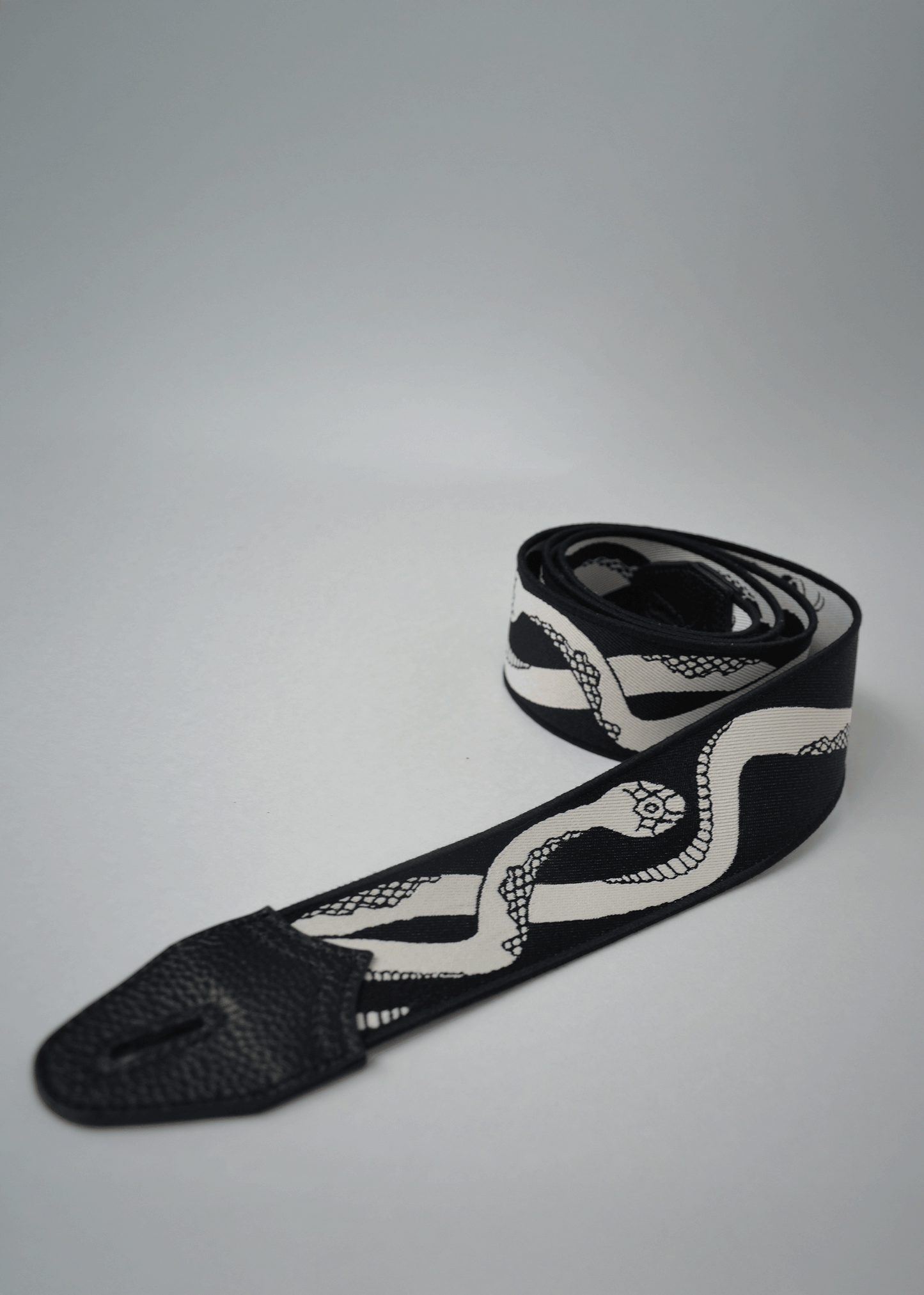 Rolled black side view of reversible snake jacquard guitar strap with black leather endcap by Mary and Shelley.
