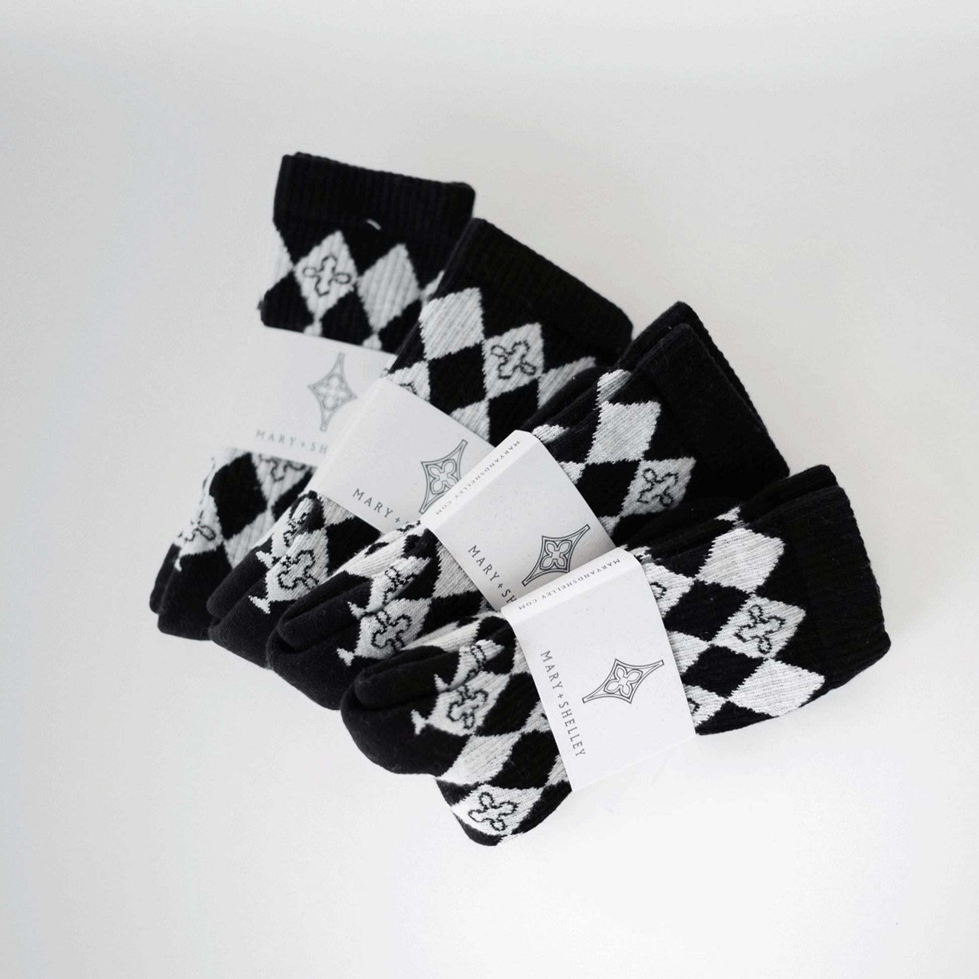 4 pairs of black and light gray patterned crew socks with a harlequin diamond design and small quatrefoil motifs fanned out on top of one another with belly band branded packaging attached.