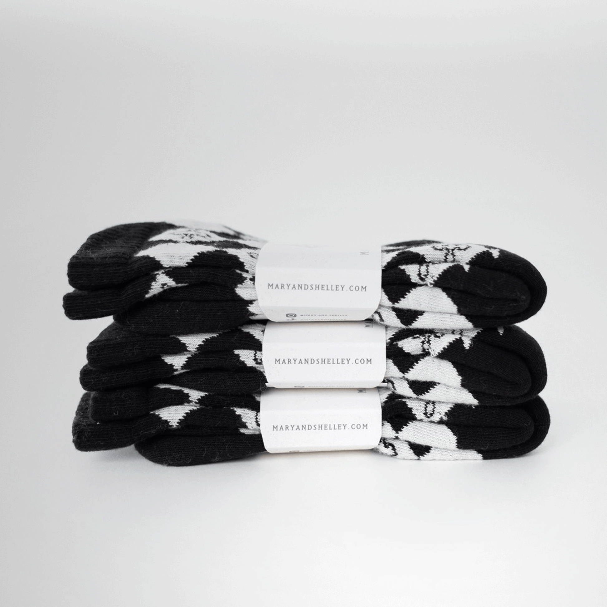 3 pairs of black and light gray patterned crew socks with a harlequin diamond design and small quatrefoil motifs stacked on top of one another with belly band branded packaging attached.