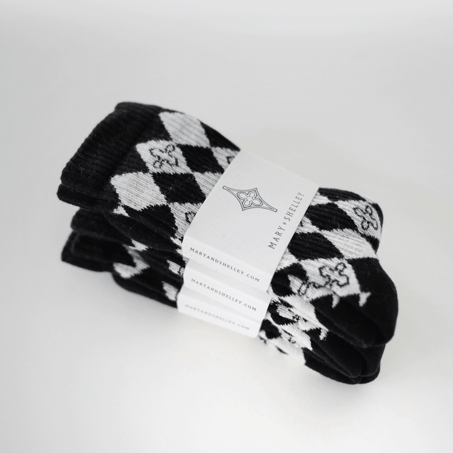 3 pairs of black and light gray patterned crew socks with a harlequin diamond design and small quatrefoil motifs stacked on top of one another with belly band branded packaging attached. Arranged at an angle.
