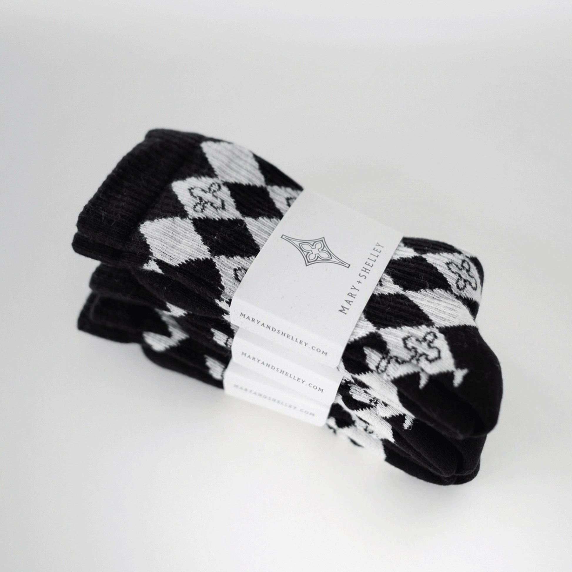 3 pairs of black and light gray patterned crew socks with a harlequin diamond design and small quatrefoil motifs stacked on top of one another with belly band branded packaging attached. Arranged at an angle.