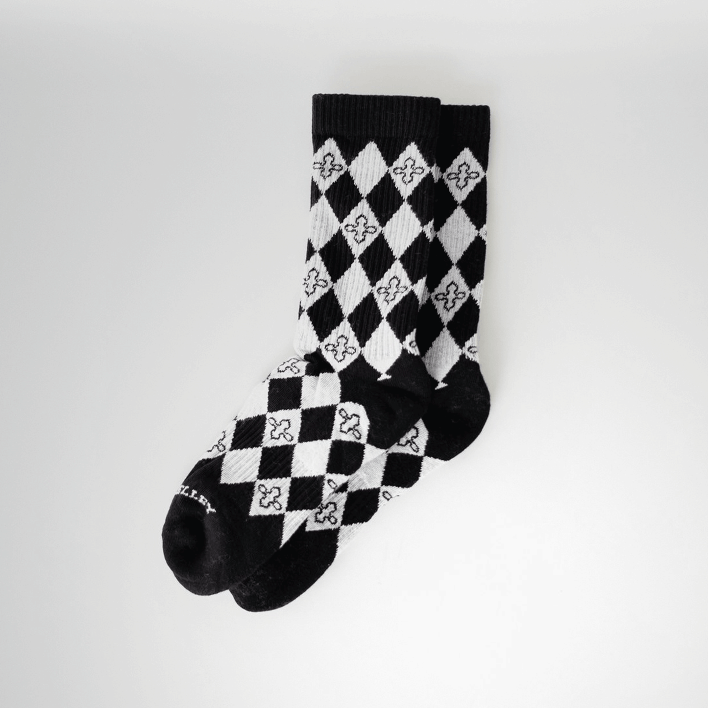 A pair of black and light gray patterned crew socks with a harlequin diamond design and small quatrefoil motifs. Laid flat, staggered on top of one another with a light gray background behind