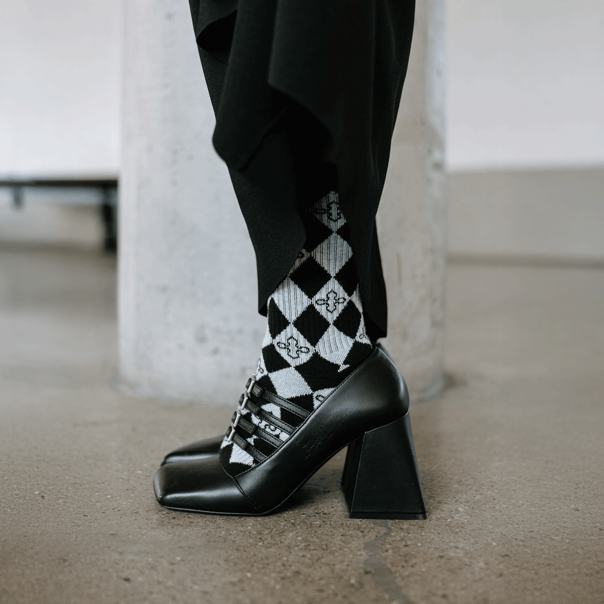 A person wearing black pointed-toe high heels with a chunky block heel and black-and-white harlequin patterned socks, standing on a concrete floor in front of a light gray column.