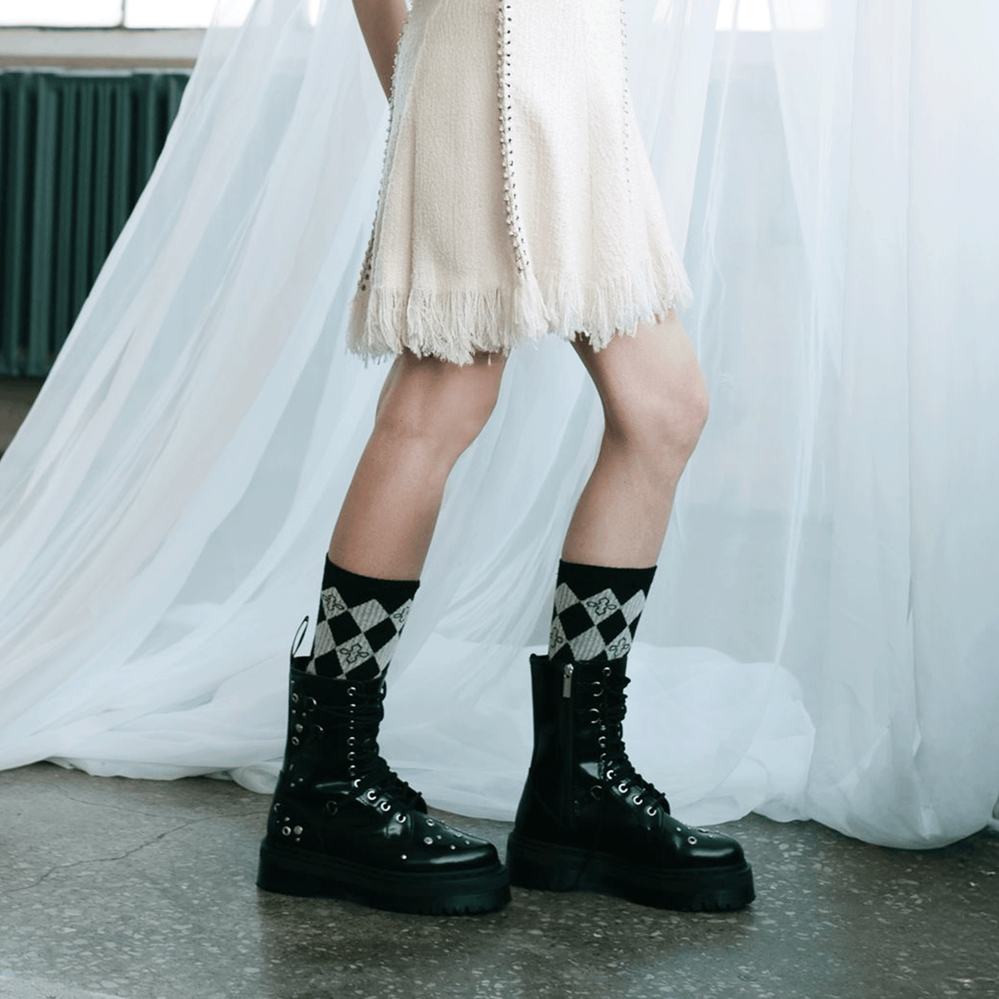 A person wearing black lace-up platform combat boots with black and grey harlequin socks, standing in front of a white draped curtain.