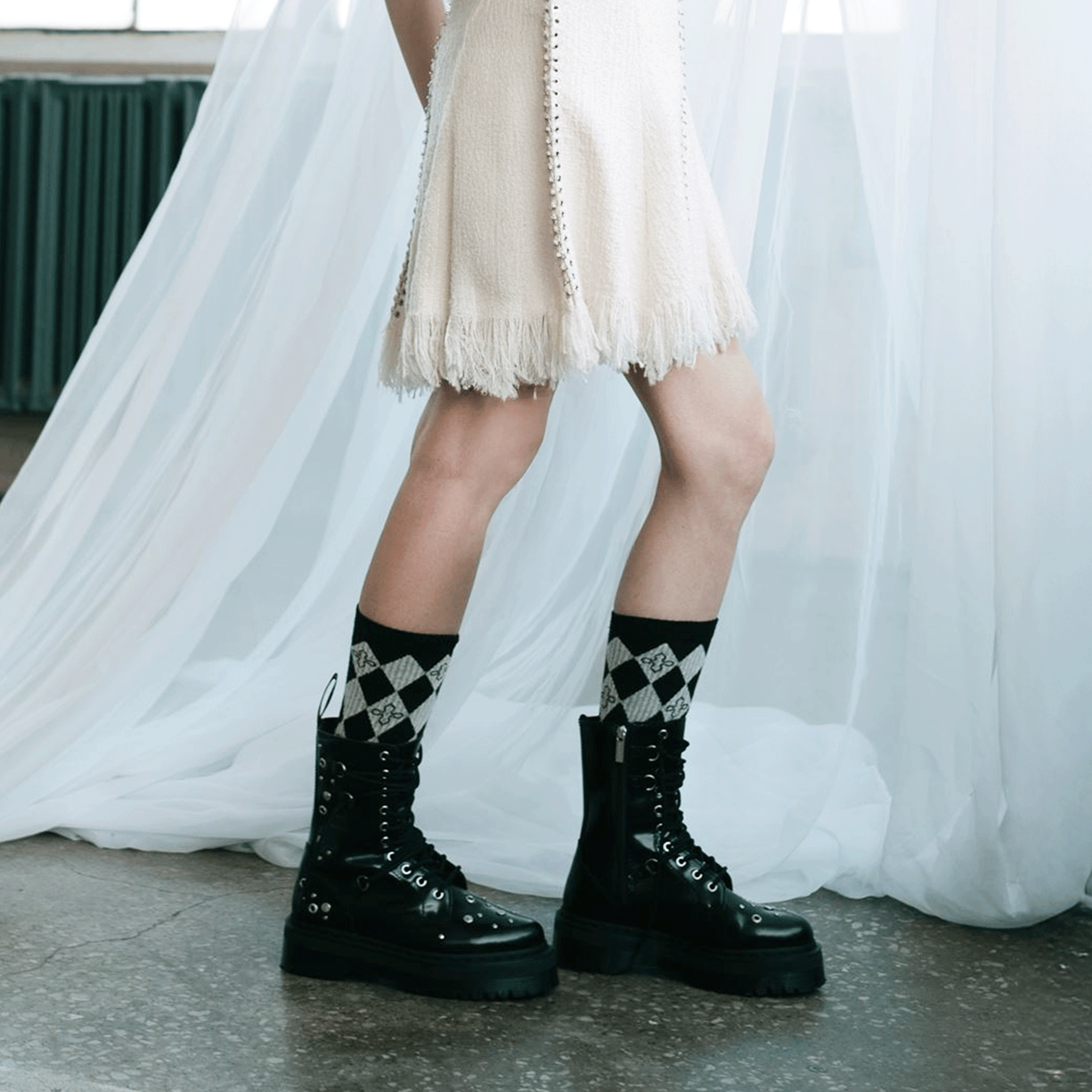 A person wearing black lace-up platform combat boots with black and grey harlequin socks, standing in front of a white draped curtain.
