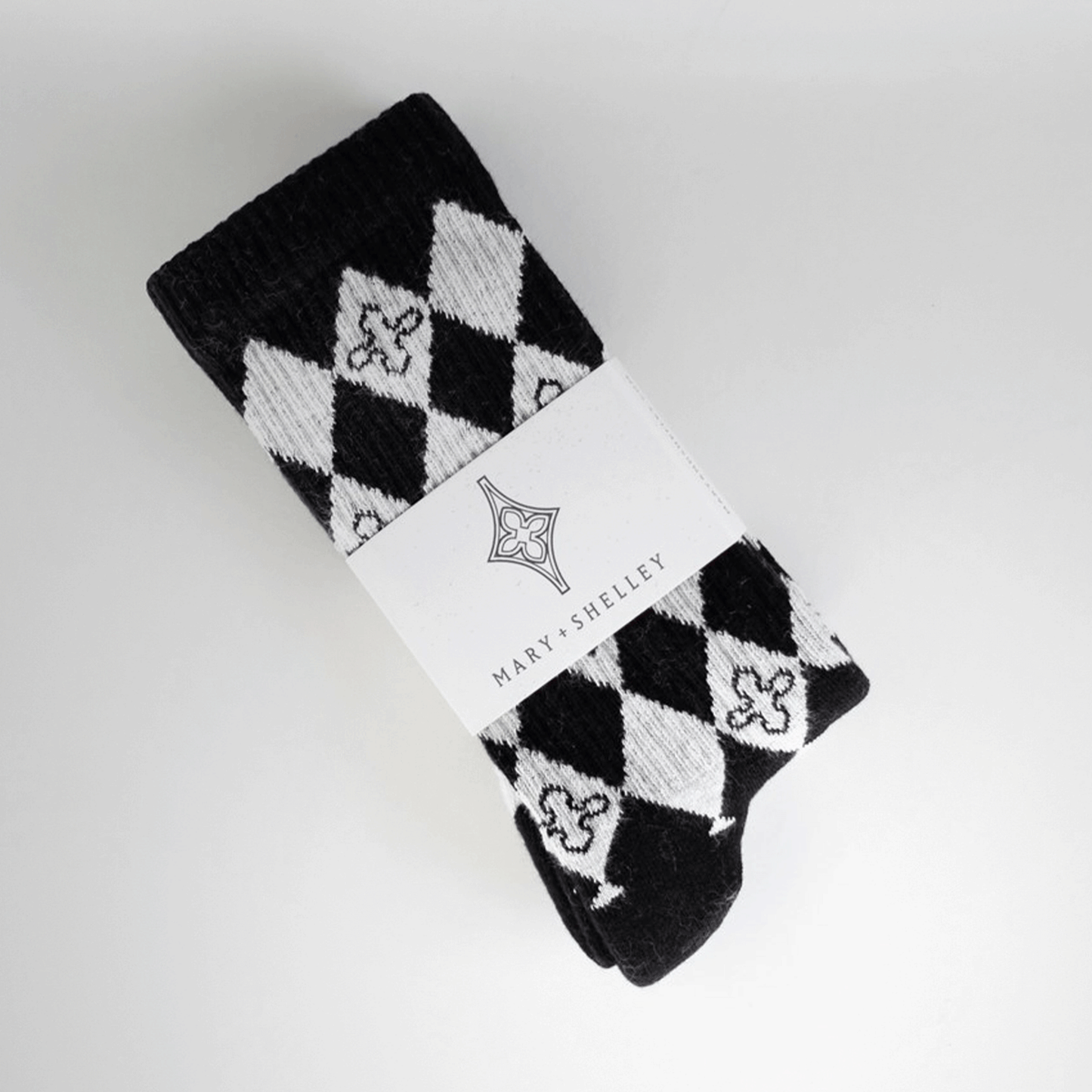 A pair of black and light gray patterned crew socks with a harlequin diamond design and small quatrefoil motifs. In belly band branded packaging, laid flat at a diagonal on a light gray background.