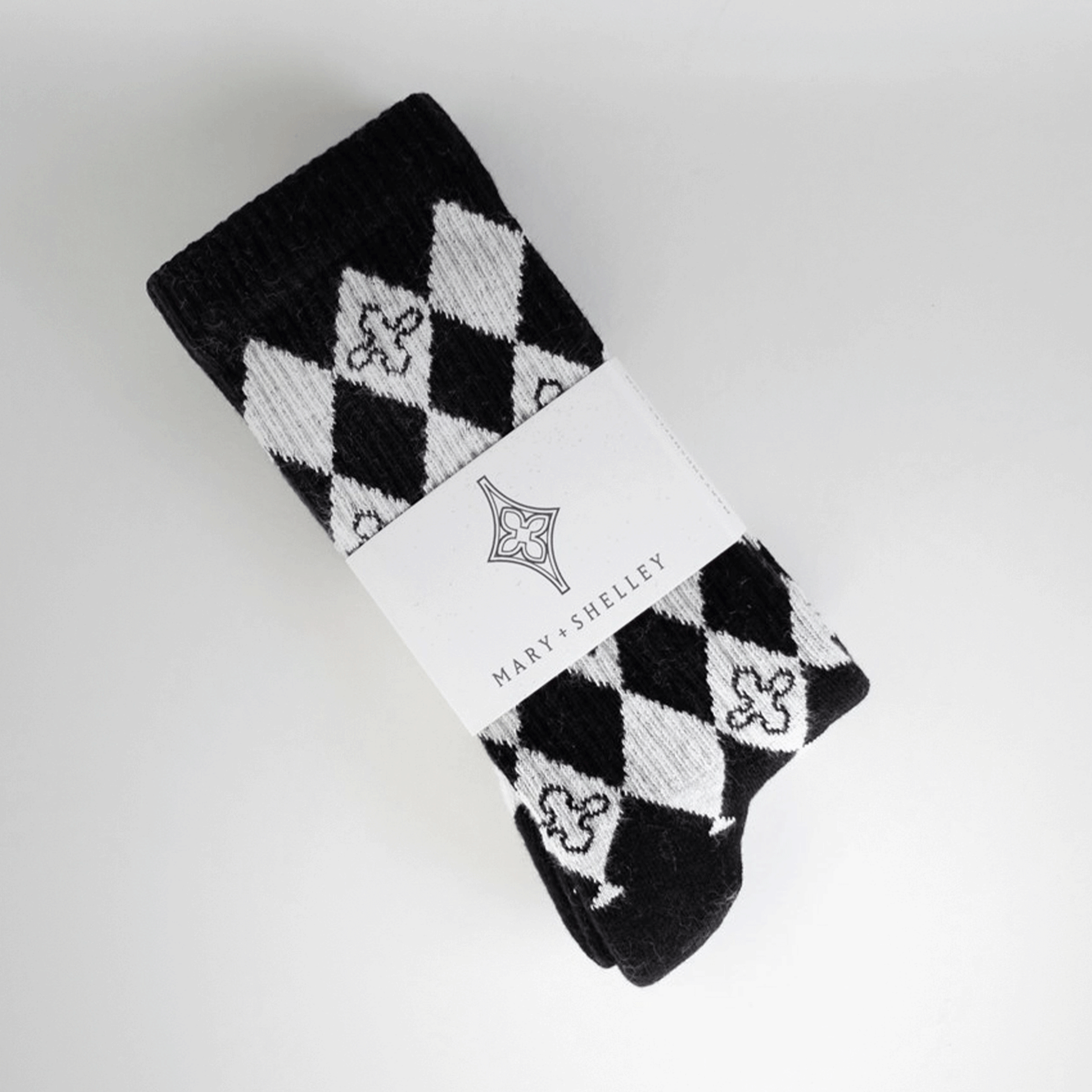 A pair of black and light gray patterned crew socks with a harlequin diamond design and small quatrefoil motifs. In belly band branded packaging, laid flat at a diagonal on a light gray background.