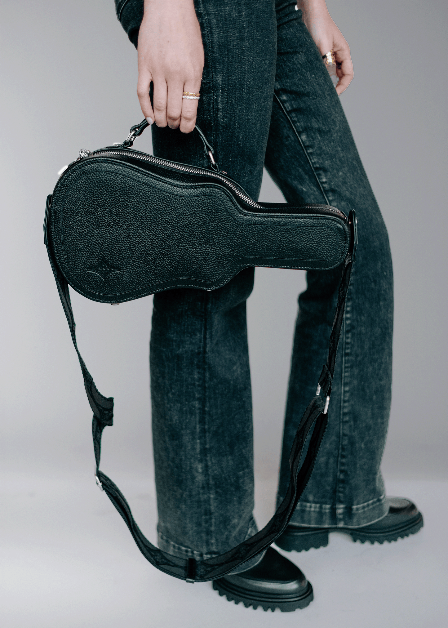 Model holding black Mary and Shelley Ritual Crossbody Bag with guitar case silhouette and jacquard logo strap, shown worn as a handheld clutch.