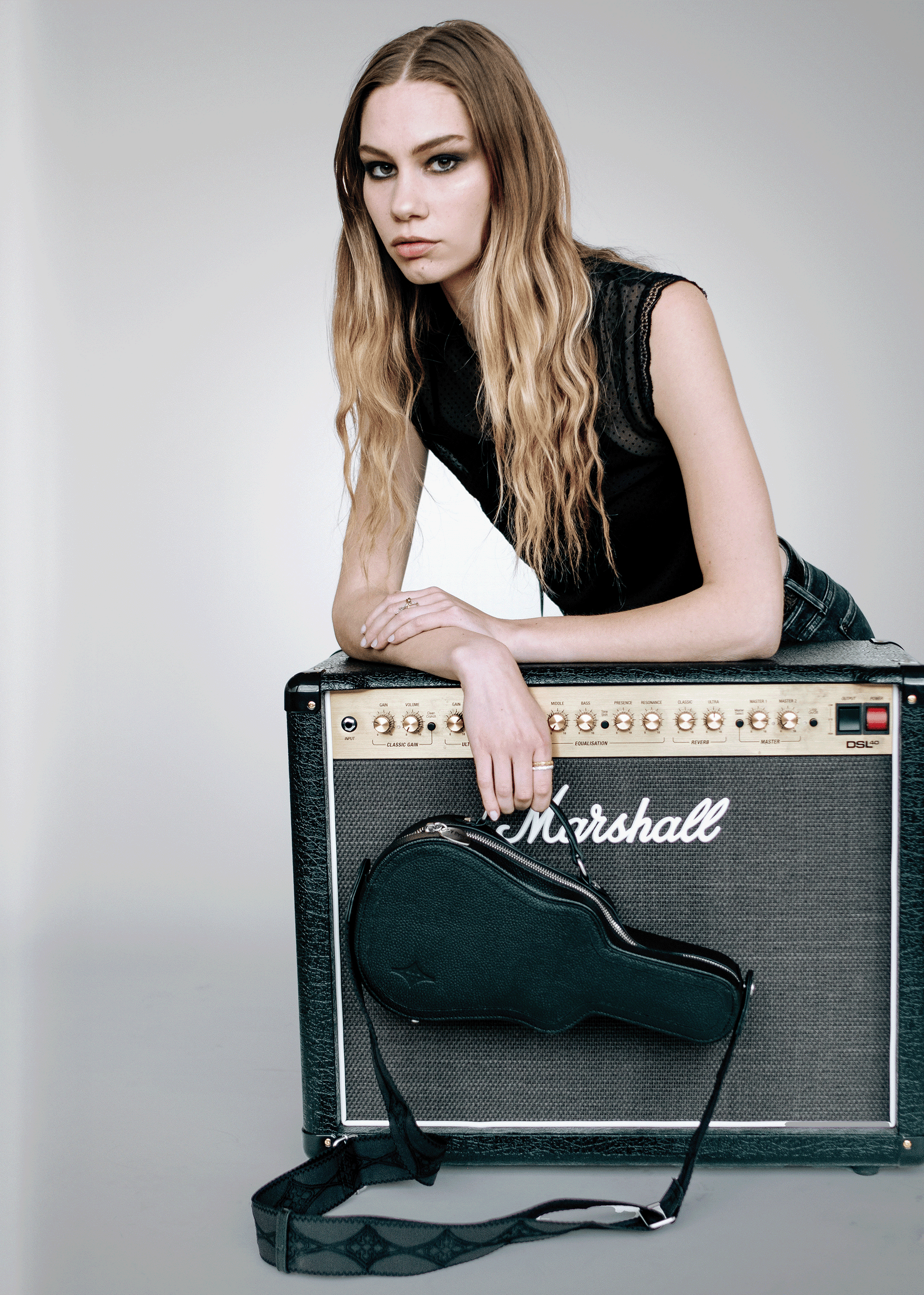 Model with black Mary and Shelley Ritual Crossbody Bag styled on a black amplifier, featuring guitar case silhouette and jacquard logo strap.