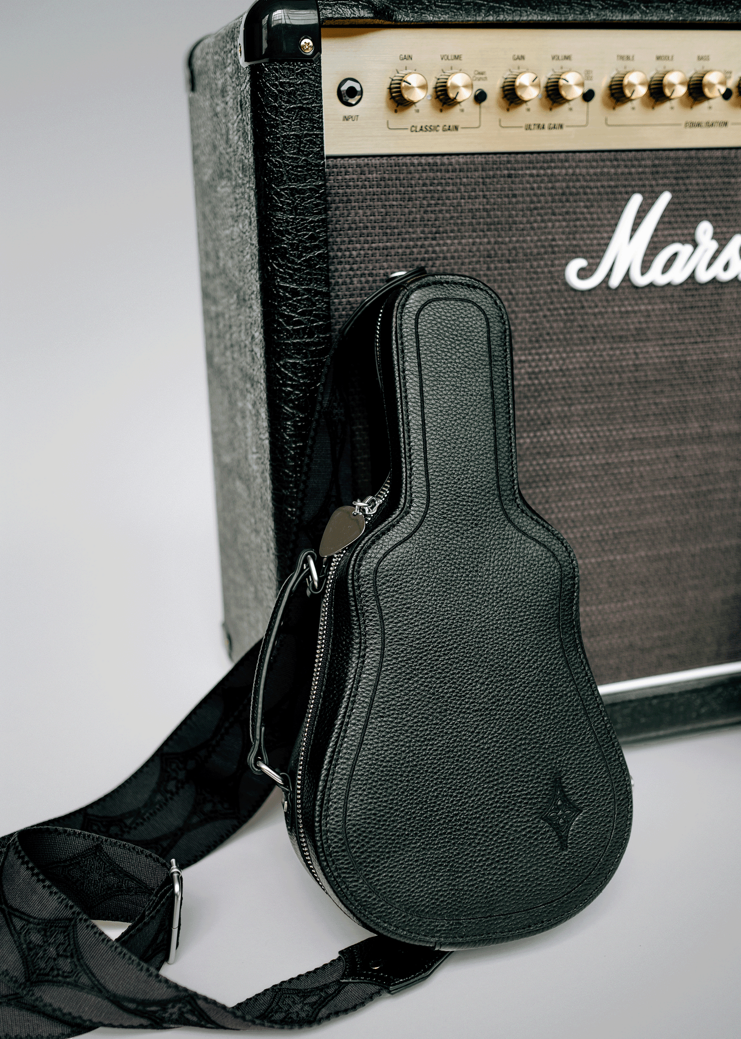 Black Mary and Shelley Ritual Crossbody Bag with guitar case silhouette, metal guitar pick zipper pull, and jacquard logo strap, styled leaning against a black amplifier.
