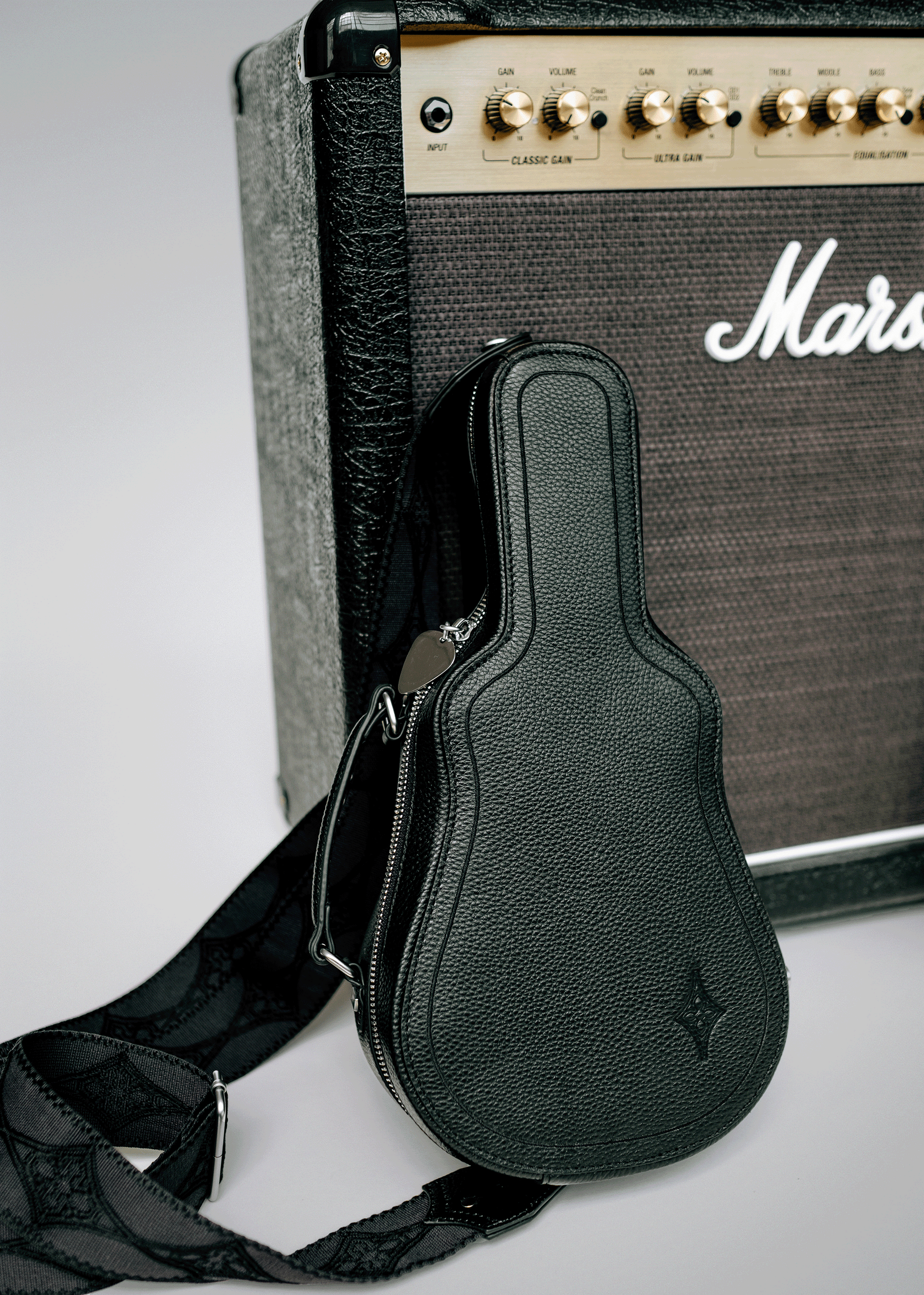 Black Mary and Shelley Ritual Crossbody Bag with guitar case silhouette, metal guitar pick zipper pull, and jacquard logo strap, styled leaning against a black amplifier.