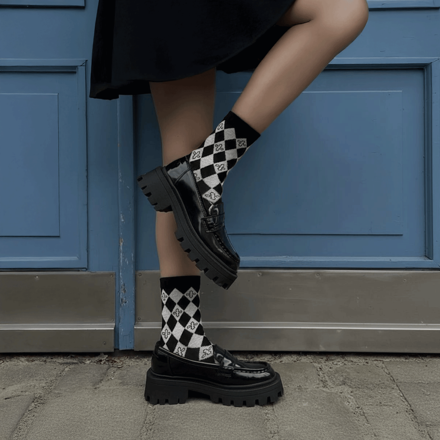 A person wearing black chunky patent leather loafers with thick platform soles and black-and-white checkered harlequin socks, standing in front of a blue paneled door.