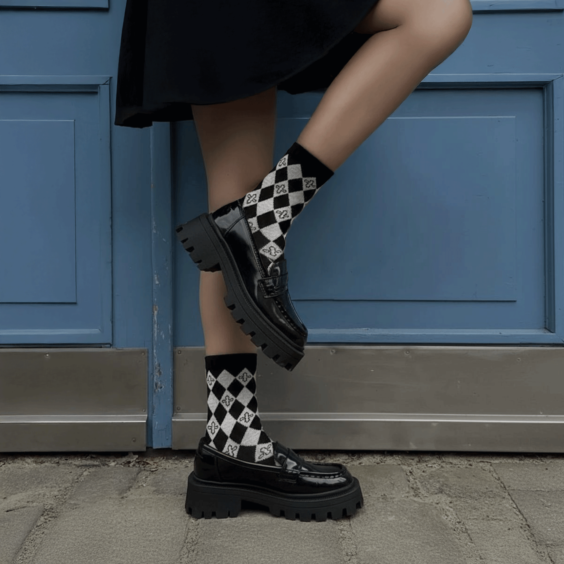 A person wearing black chunky patent leather loafers with thick platform soles and black-and-white checkered harlequin socks, standing in front of a blue paneled door.