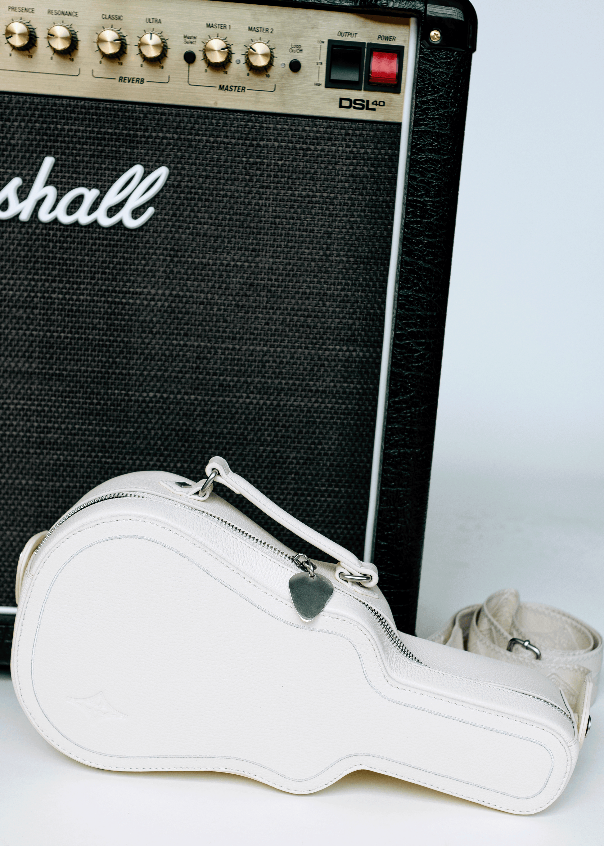 White (Ivory) Mary and Shelley Ritual Crossbody Bag with guitar case silhouette and metal guitar pick zipper pull, styled next to black amplifier.